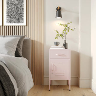 Metal side table locker in light pink with one drawer and one door, in a room next to a bed